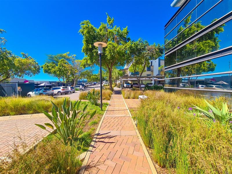 A well-maintained exterior walkway featuring brick paving, landscaped garden beds, and a modern glass facade office building.
