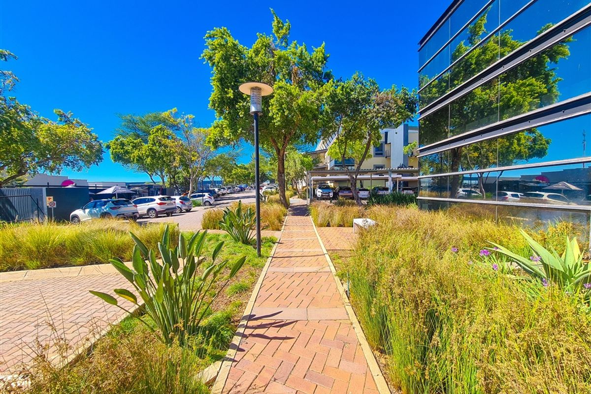 A well-maintained exterior walkway featuring brick paving, landscaped garden beds, and a modern glass facade office building.