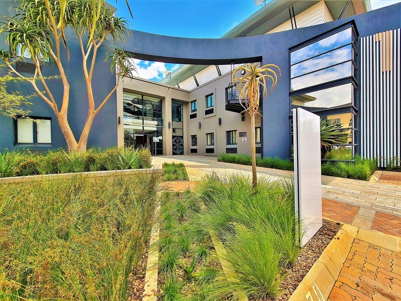 Modern office building entrance featuring contemporary architectural design, glass facade elements, and well-maintained landscaped garden beds with a paved approach.