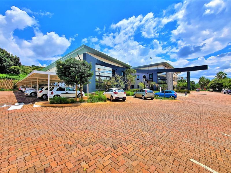 Modern office building exterior featuring a large paved parking area, covered parking bays, and contemporary architectural design with extensive glass paneling.
