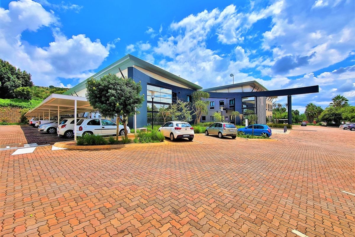 Modern office building exterior featuring a large paved parking area, covered parking bays, and contemporary architectural design with extensive glass paneling.