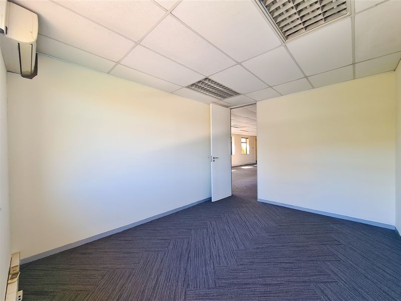 A clean, empty office space featuring carpet tiles, a suspended ceiling with recessed lighting, and a wall-mounted air conditioning unit.