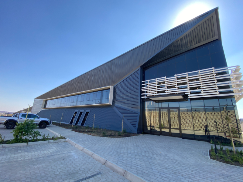 Modern industrial building facade featuring dark metal cladding, extensive glass curtain walling, and a paved entrance area.