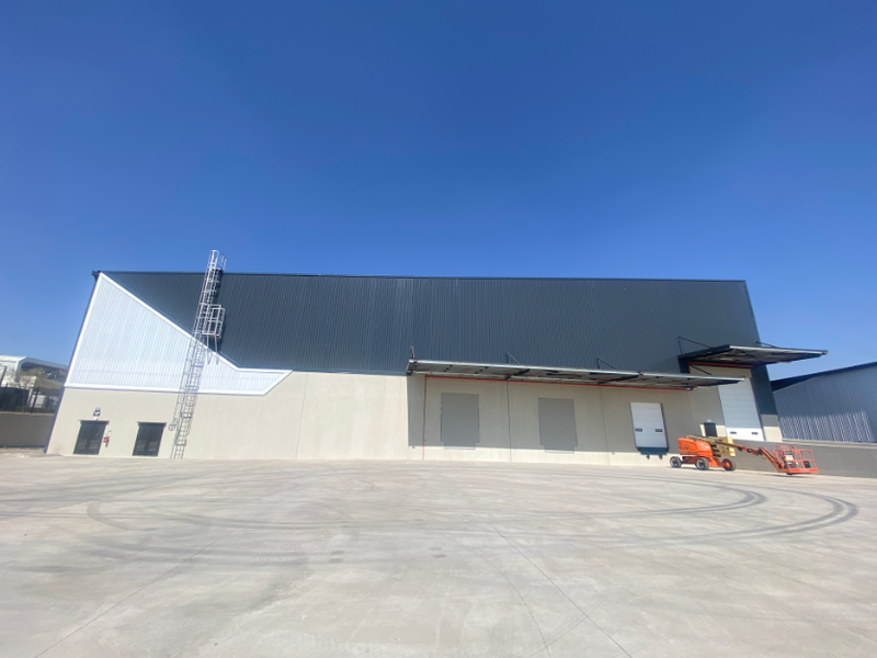 Modern industrial warehouse exterior featuring large metal cladding, multiple roller shutter loading doors, a concrete hardstand area, and an external access ladder.