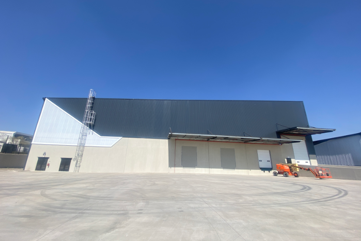 Modern industrial warehouse exterior featuring large metal cladding, multiple roller shutter loading doors, a concrete hardstand area, and an external access ladder.