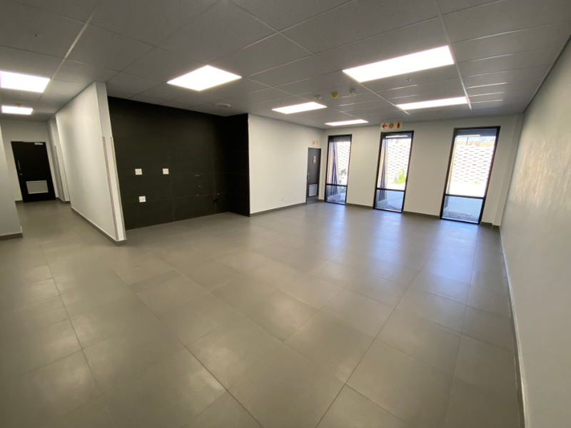 A spacious, modern, and empty office area featuring neutral tiled flooring, recessed ceiling lighting, and large windows providing ample natural light.