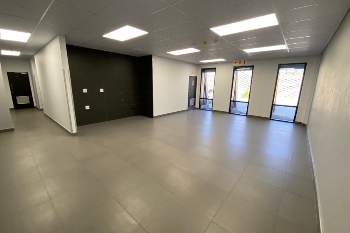A spacious, modern, and empty office area featuring neutral tiled flooring, recessed ceiling lighting, and large windows providing ample natural light.