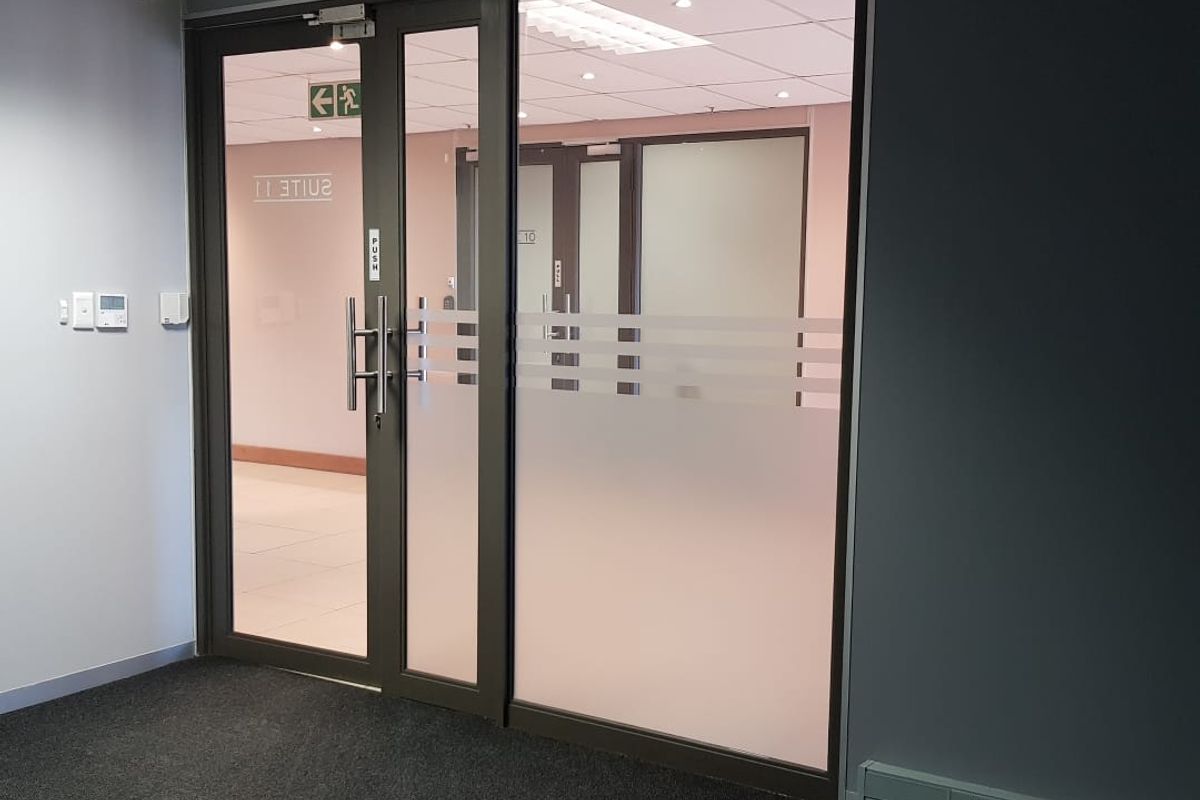 A modern office entrance featuring a glass door with frosted privacy film, suspended ceiling, and integrated electrical trunking in a professional commercial setting.