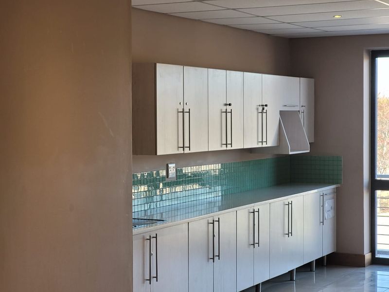 A clean, functional office kitchenette featuring light wood-finish cabinetry, a green tiled backsplash, and integrated sink area.