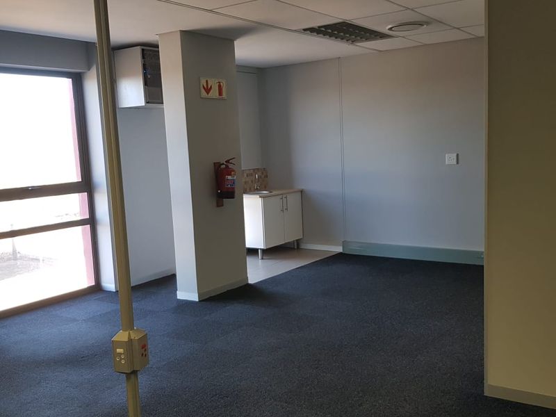 An open-plan office space featuring carpeted flooring, a suspended ceiling, a small kitchenette area with a sink, and a central power pole.