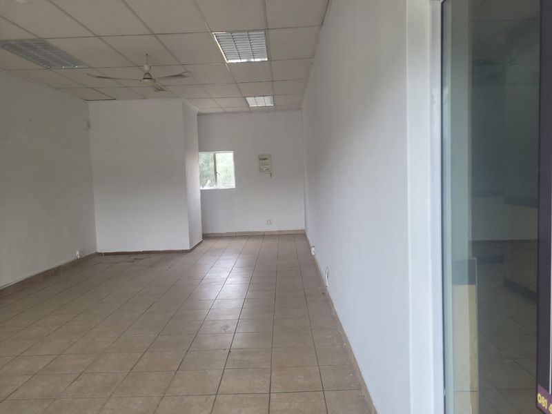 An empty, open-plan retail space featuring tiled flooring, a suspended ceiling with recessed lighting, and a large glass storefront entrance.
