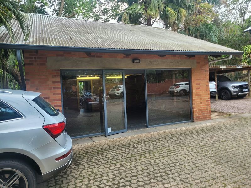 A detached office or garage space featuring exposed brickwork, a corrugated metal roof, and modern aluminium-framed glass sliding doors, set within a paved driveway area.