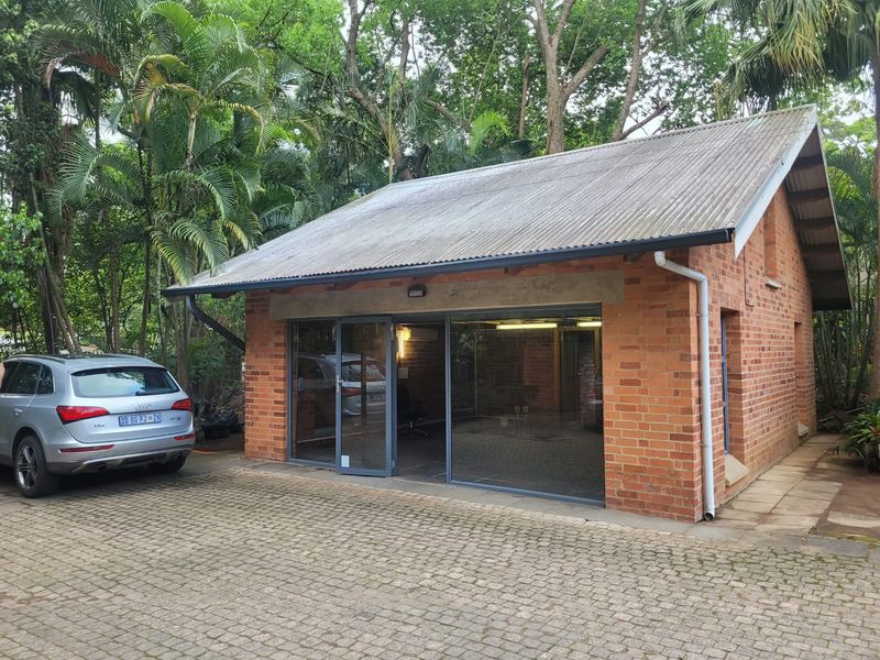 A detached office building featuring a classic brick facade, corrugated metal roof, and modern glass sliding doors, set within a lush, tree-lined environment.
