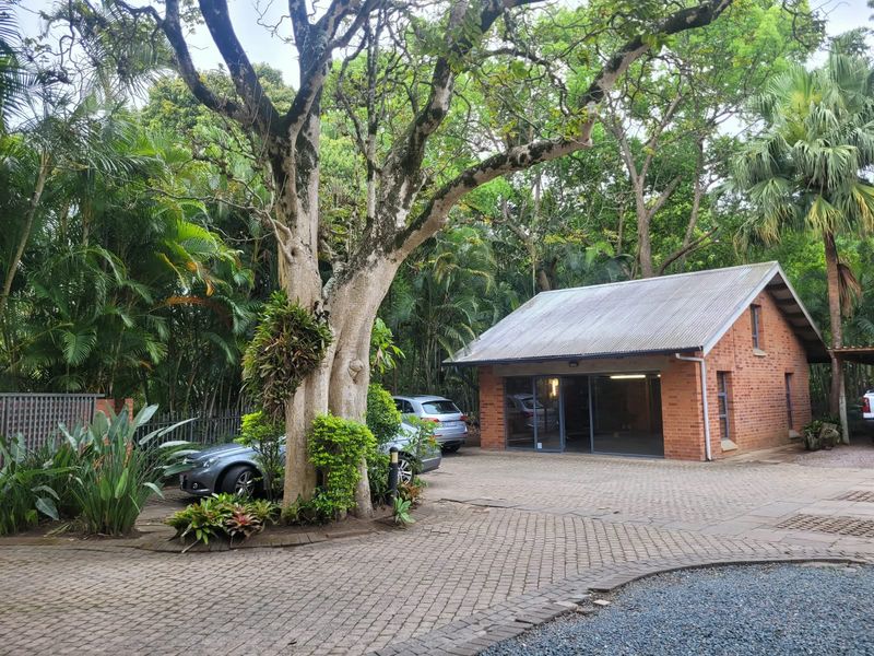 A well-maintained brick office building with a corrugated metal roof, large glass sliding doors, and a paved driveway set in a lush garden.