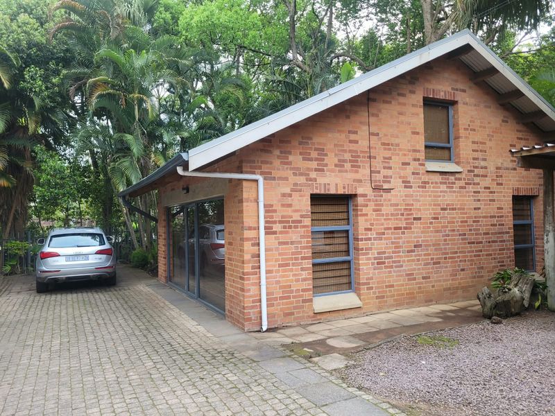 A well-maintained brick office building featuring modern aluminium window frames, a pitched roof, and a paved driveway set within a lush, green garden environment.
