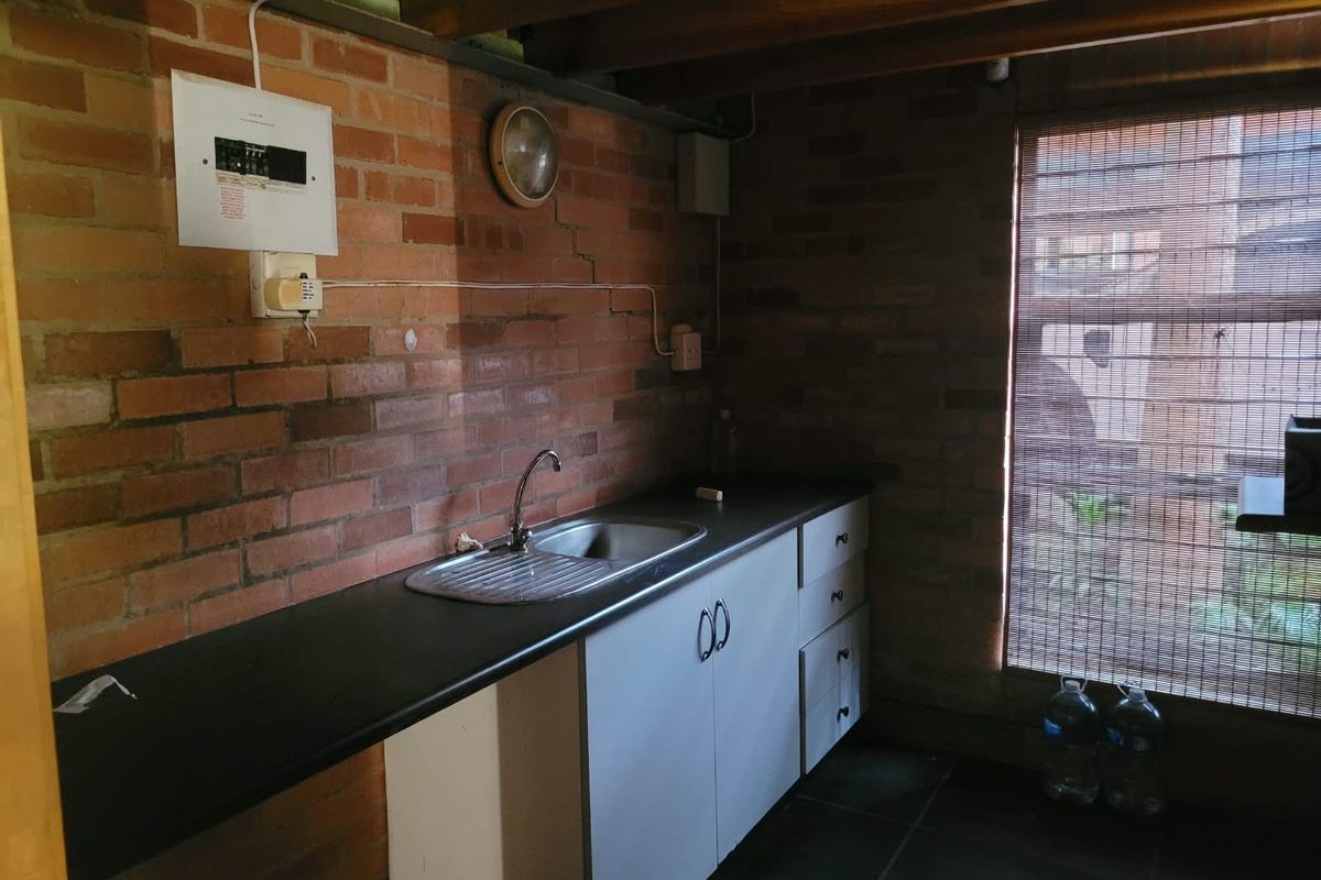A compact kitchenette featuring exposed brick walls, timber ceiling beams, built-in cabinetry, and a stainless steel sink, requiring some maintenance to the brickwork.