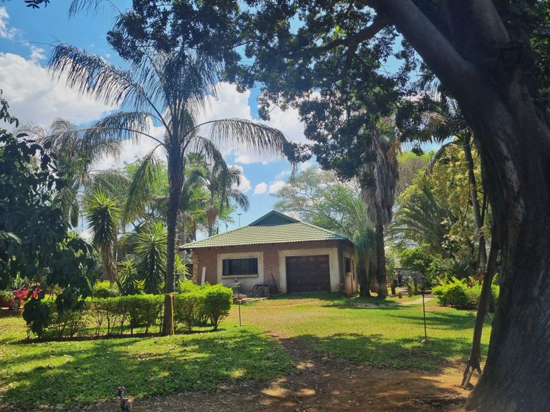 A view of a brick-built outbuilding or garage set within a mature, landscaped garden on a farm property.