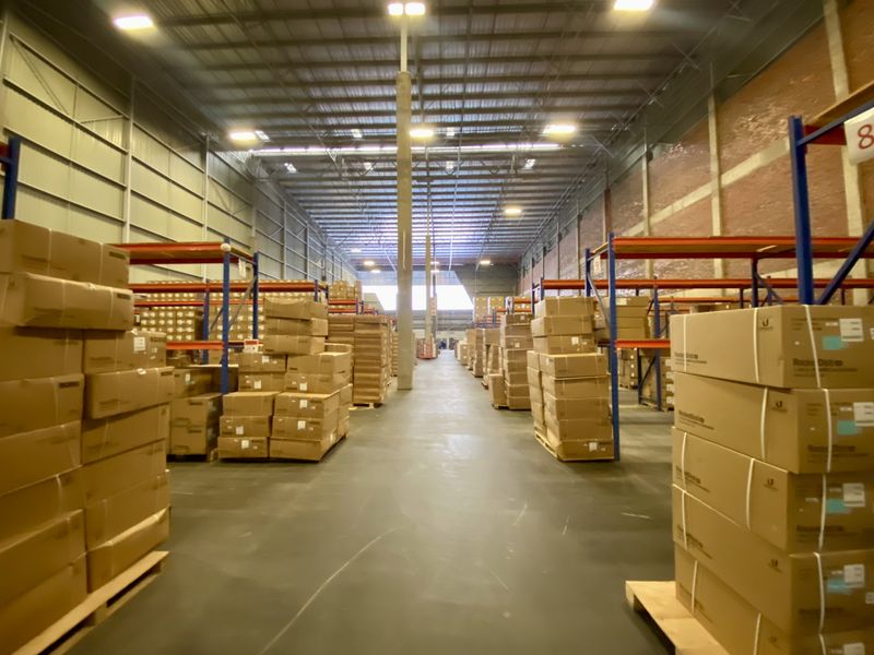 Spacious industrial warehouse featuring high-bay lighting, exposed steel structural beams, concrete flooring, and extensive heavy-duty racking systems for storage.