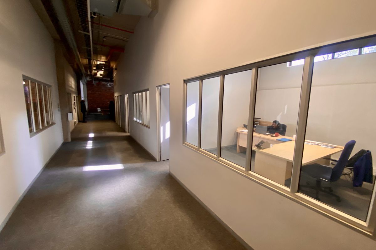 Industrial office hallway featuring exposed ceiling ductwork, carpeted flooring, and glass partition walls looking into private office spaces.
