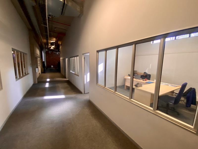 Industrial office hallway featuring exposed ceiling ductwork, carpeted flooring, and glass partition walls looking into private office spaces.