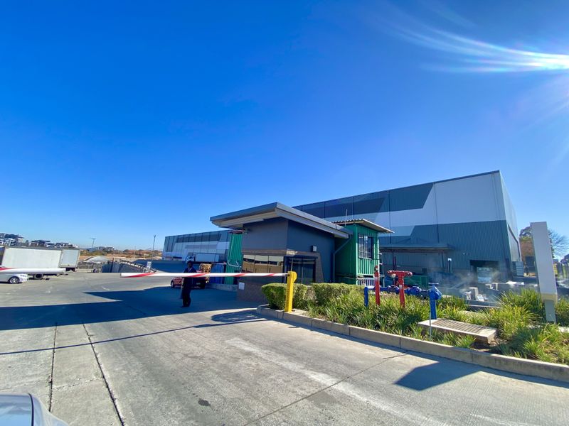 Industrial property entrance featuring a security boom gate, guard house, and large warehouse facility with paved access and landscaped surroundings.
