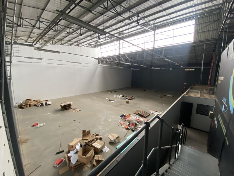 Spacious industrial warehouse unit featuring high ceilings with exposed steel trusses, concrete flooring, mezzanine access, and ample natural light from overhead skylights.