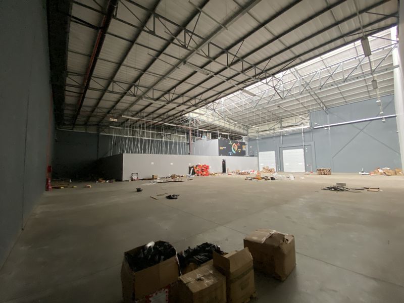 Spacious industrial warehouse featuring high ceilings with exposed steel trusses, concrete flooring, large roller shutter doors, and natural light from overhead skylights.