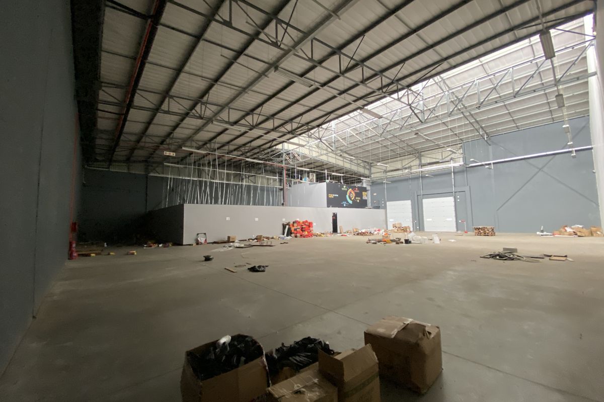 Spacious industrial warehouse featuring high ceilings with exposed steel trusses, concrete flooring, large roller shutter doors, and natural light from overhead skylights.
