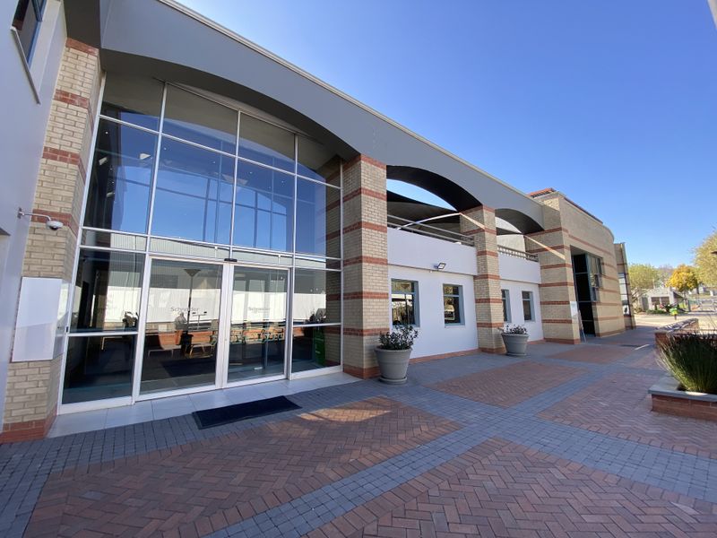 Modern industrial office building entrance featuring a large glass curtain wall, brick facade detailing, and a paved pedestrian courtyard area.