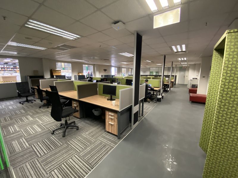 Open-plan industrial office space featuring modular cubicle workstations, suspended ceilings with recessed lighting, and a mix of carpeted and smooth flooring.