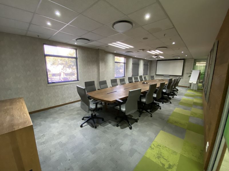 A professional boardroom featuring a large central table, suspended ceiling with recessed lighting, carpet tile flooring, and a wall-mounted projector screen.