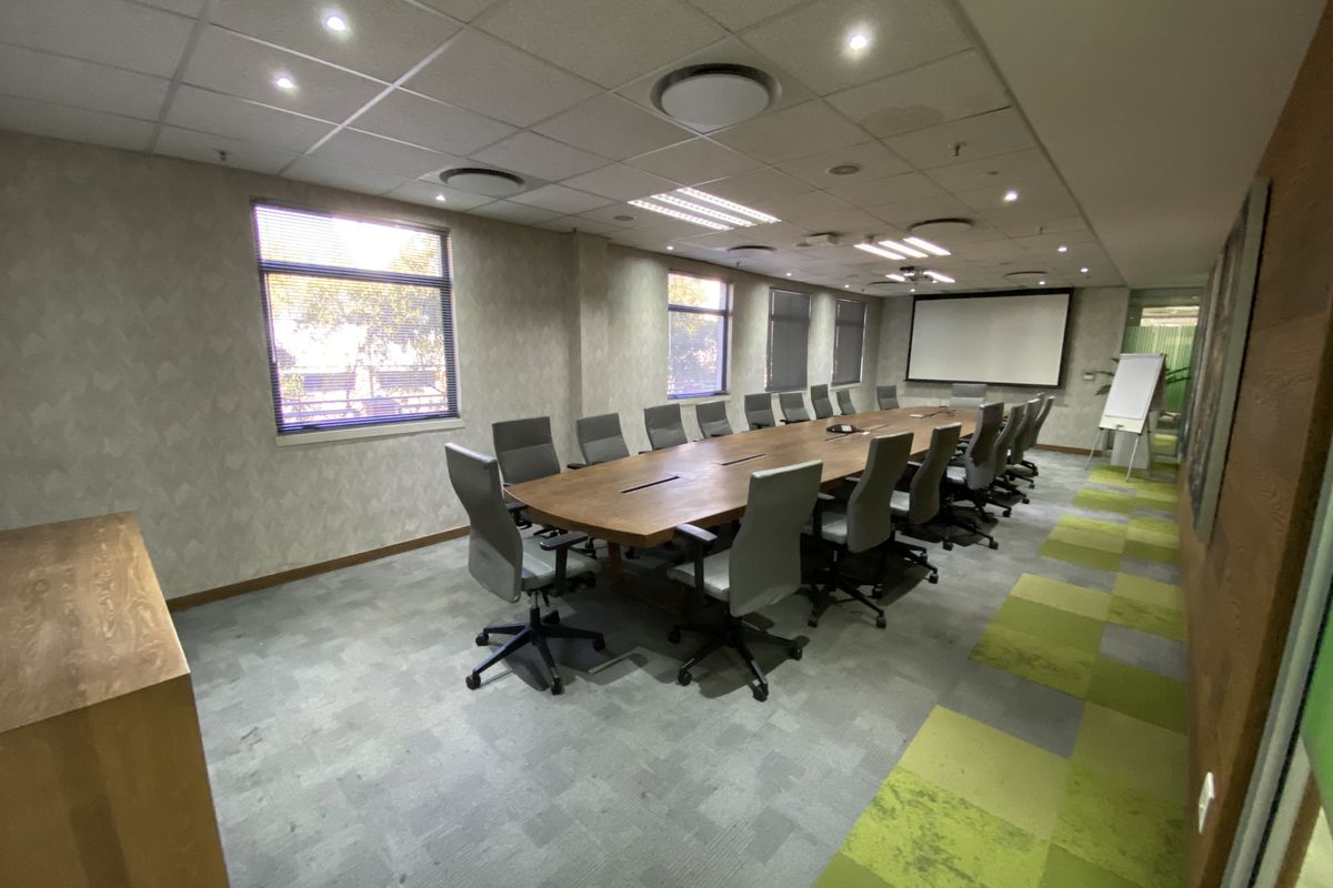 A professional boardroom featuring a large central table, suspended ceiling with recessed lighting, carpet tile flooring, and a wall-mounted projector screen.