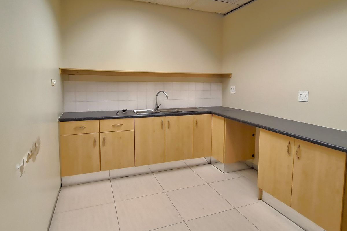 A functional office kitchenette featuring light wood cabinetry, dark laminate countertops, a stainless steel sink, and tiled flooring.