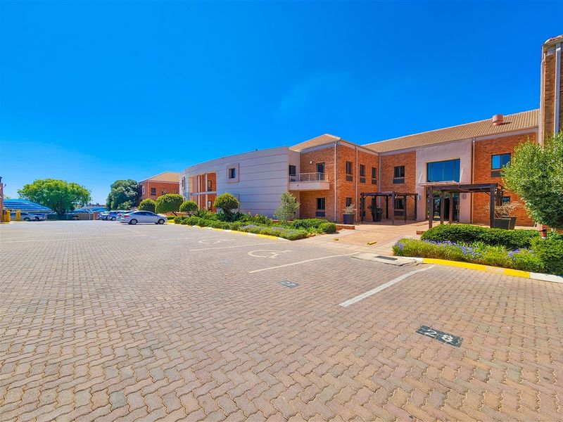 A well-maintained office building exterior featuring a large paved parking area, brick facade, and landscaped garden beds at the entrance.