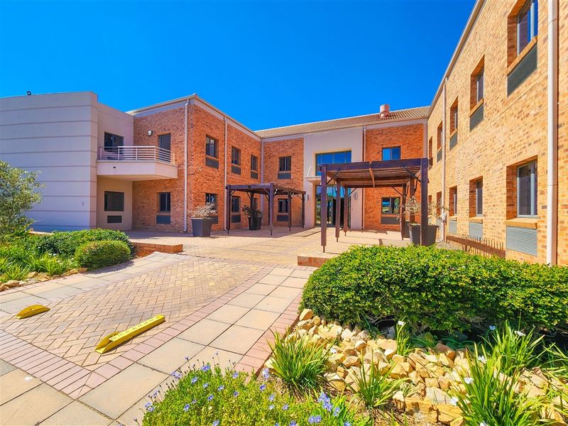 A well-maintained office building exterior featuring a brick facade, paved courtyard area with timber pergolas, and landscaped garden beds.
