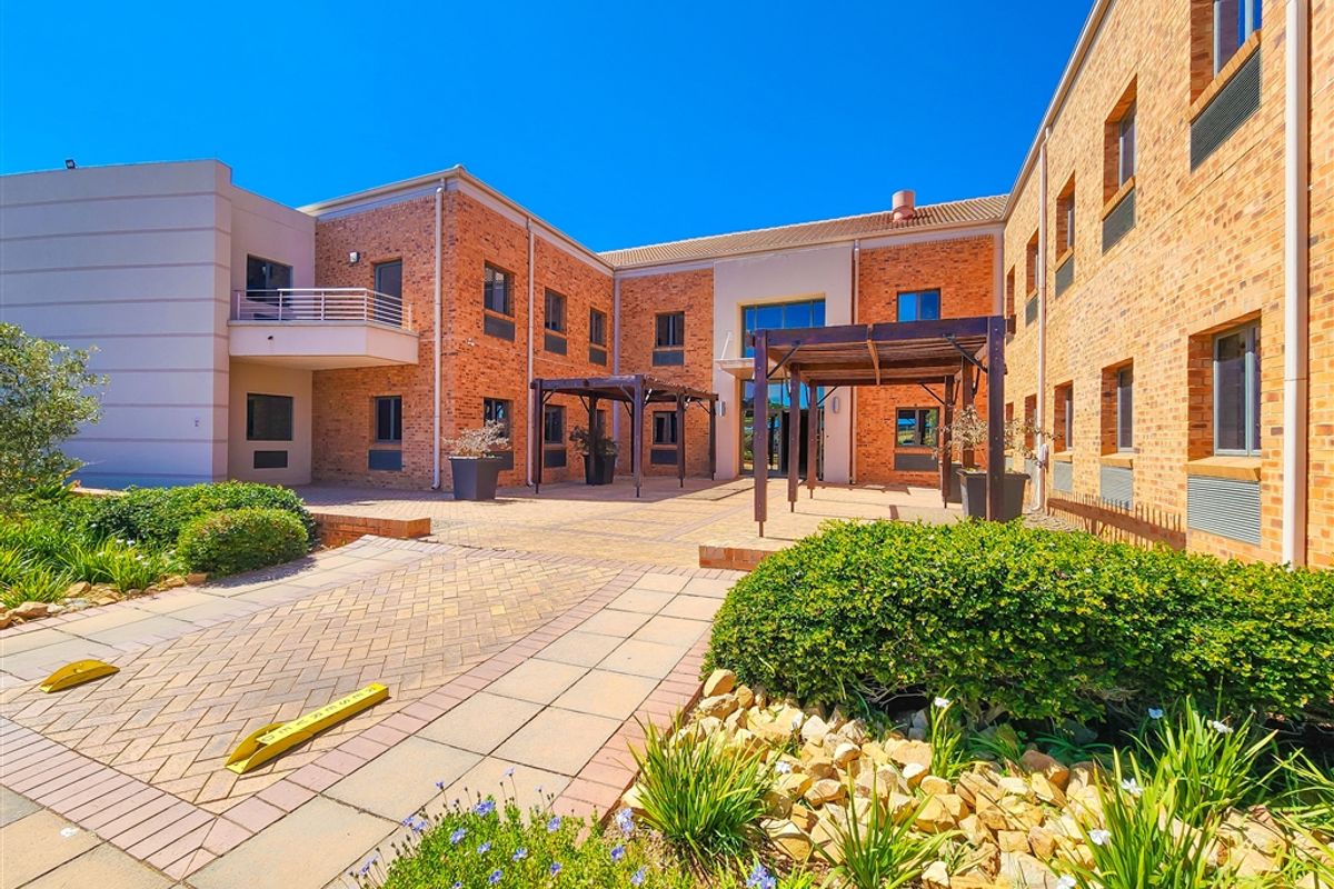 A well-maintained office building exterior featuring a brick facade, paved courtyard area with timber pergolas, and landscaped garden beds.