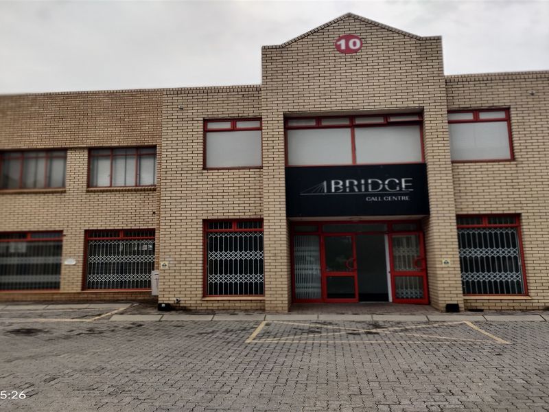 Front elevation of a commercial industrial building featuring a brick facade, paved driveway, and secure entrance with red-framed windows and security gates.