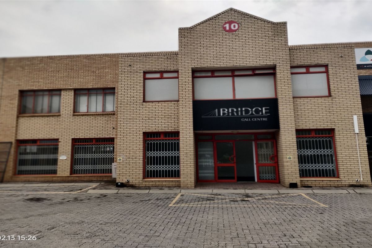 Front elevation of a commercial industrial building featuring a brick facade, paved driveway, and secure entrance with red-framed windows and security gates.