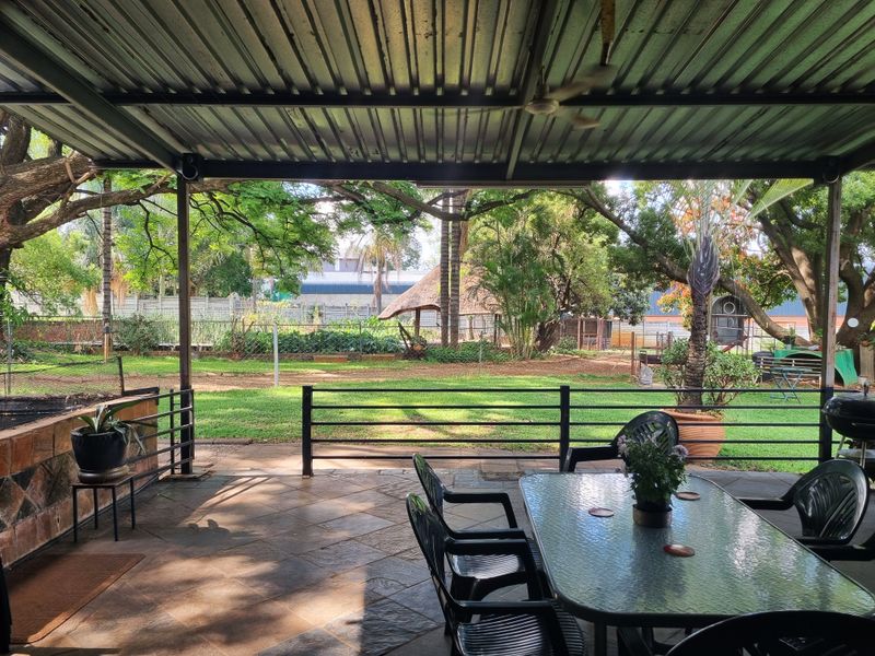 A covered outdoor patio featuring tiled flooring, a metal roof, and a built-in brick braai, overlooking a spacious garden area.