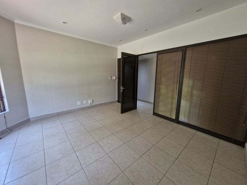 A bright, empty office space featuring neutral tiled flooring, recessed lighting, and large aluminium-framed glass doors with integrated venetian blinds.