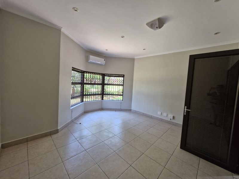 A bright, empty office space featuring tiled flooring, a bay window with security bars, and a wall-mounted air conditioning unit.
