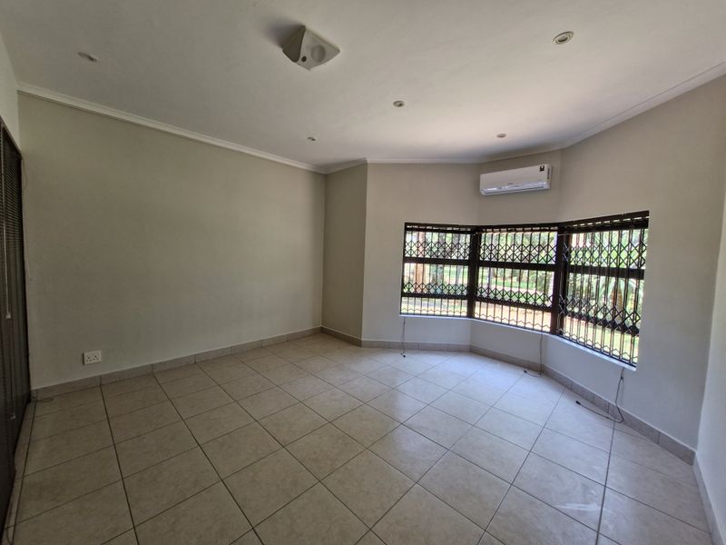 An empty, well-lit living area featuring tiled flooring, a bay window with security bars, and a wall-mounted air conditioning unit.