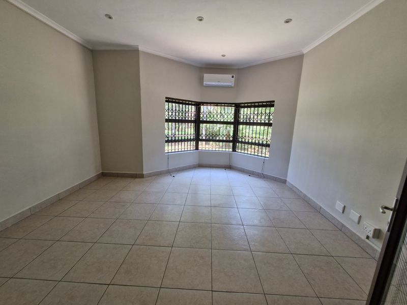 A bright, empty living room featuring neutral tiled flooring, a bay window with security gates, and a wall-mounted air conditioning unit.
