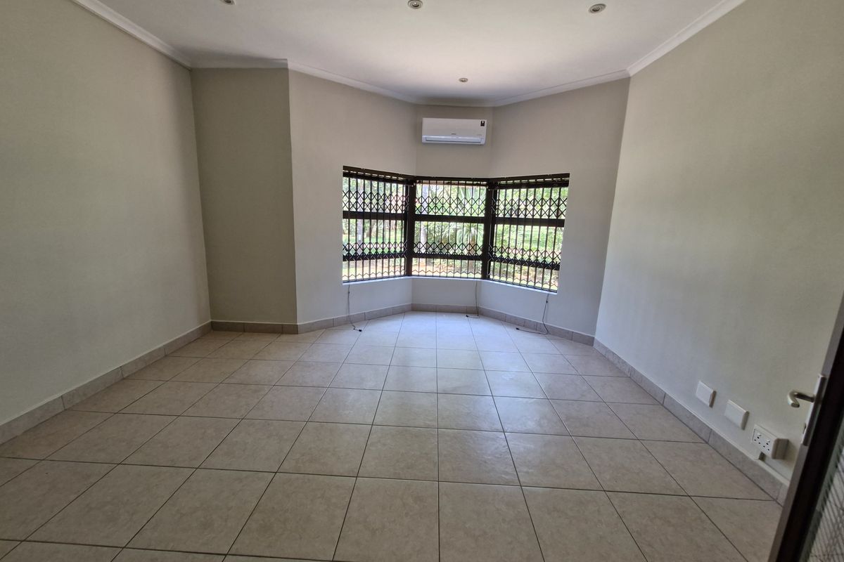 A bright, empty living room featuring neutral tiled flooring, a bay window with security gates, and a wall-mounted air conditioning unit.