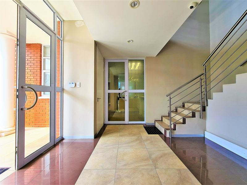 A bright and modern office building entrance featuring tiled flooring, a stainless steel staircase railing, and glass-paneled doors leading to the interior.