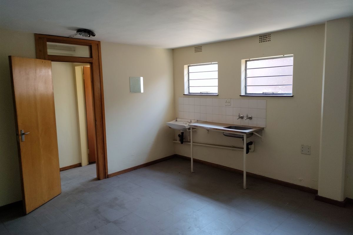 A basic, empty utility kitchen area featuring a stainless steel sink, tiled splashback, and simple metal-framed windows in an industrial property setting.