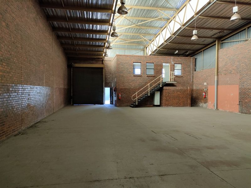 Spacious industrial warehouse unit featuring high ceilings with exposed steel trusses, concrete flooring, a roller shutter door, and an internal office mezzanine level.