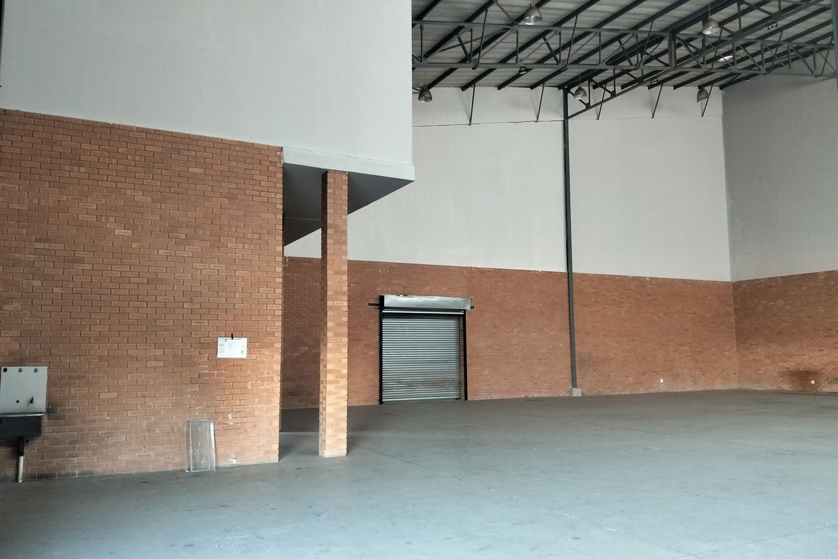 Spacious industrial warehouse unit featuring high ceilings with exposed steel trusses, concrete flooring, brick walls, and a secure roller shutter door.