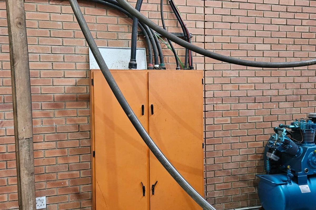 Industrial utility area featuring an electrical distribution cabinet mounted on an exposed brick wall with associated industrial air compressor and exposed conduit piping.