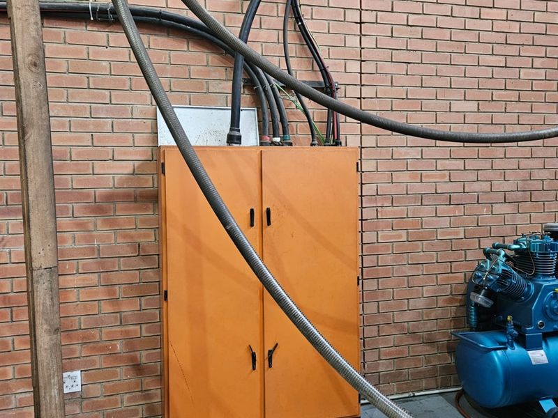 Industrial utility area featuring an electrical distribution cabinet mounted on an exposed brick wall with associated industrial air compressor and exposed conduit piping.